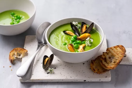 Bol blanc rempli de gaspacho vert avec des moules, petits pois et fleurs comestibles à la surface.
