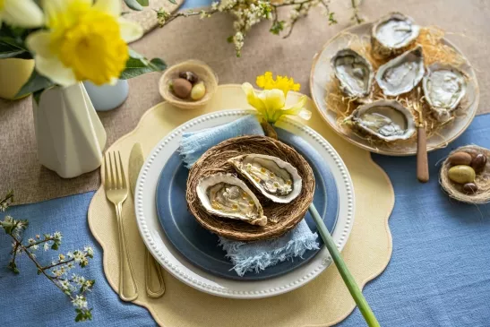 Une personne décrochant une huître de sa coquille lors d'une dégustation, la scène se déroule sur une table de pique-nique en bois sur laquelle est disposée une assiette de douze huîtres et un verre de vin blanc.
