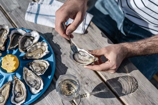 Une personne décrochant une huître de sa coquille lors d'une dégustation, la scène se déroule sur une table de pique-nique en bois sur laquelle est disposée une assiette de douze huîtres et un verre de vin blanc.