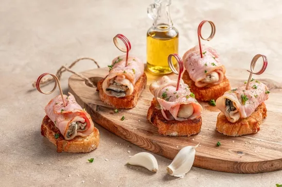 Planche à découper en bois sur laquelle est disposée des tartines d'huîtres au bacon. Une recette conviviale qui sent le partage.